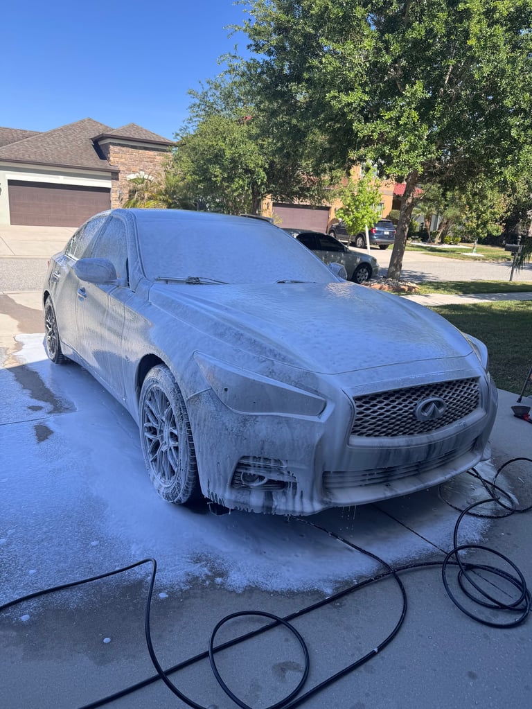 Blue Infiniti sedan covered in soapy water being washed in a residential driveway with hoses visible